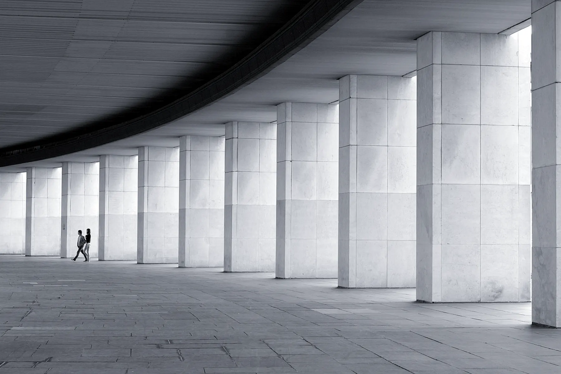 A vast bone-white colonnade in cool light; two figures walking give it human scale.